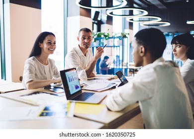 Smiling man and woman looking at coworkers and talking about startup project while sitting at table in boardroom in office