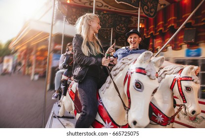 Smiling Man And Woman On Amusement Park Carousel. Young Couple On Horse Carousel Ride At Fairground.