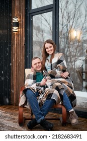 Smiling Man And Woman Wrapped In Blanket Sitting In Chair Outside Scandinavian House Barnhouse. Happy Couple In Love Resting Together Outdoors Near Building With Panoramic Window Under Winter Snow.