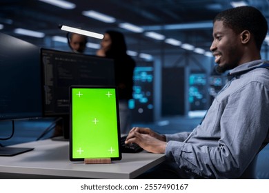 Smiling man working on green screen tablet in modern data center. Happy IT specialist doing IT infrastructure management on server racks in high tech environment using chroma key device - Powered by Shutterstock