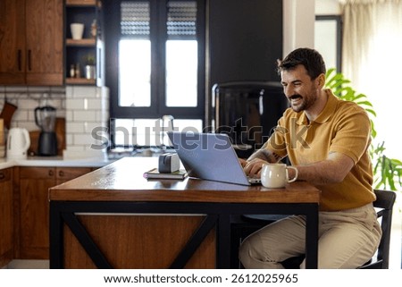 Smiling man working remotely using laptop in kitchen, enjoying flexible work schedule and work-life balance