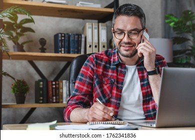 Smiling man writing notes while making phone call and using laptop at home