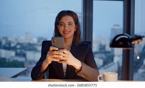 Smiling manager browsing app receiving salary at office lamp light closeup. Relaxed businesswoman typing cellphone message sitting evening workplace. Confident woman employee chatting in social media.