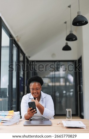 Smiling manager using her smartphone during a work break in a modern office, sitting at her organized desk with a laptop and paperwork, enjoying a moment of connection and relaxation