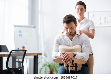 Smiling masseuse massaging shoulders of businessman sitting on massage chair in office on blurred background