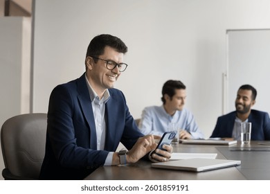 Smiling mature 45s businessman in suit sit at desk using smartphone, check updates, using new AI business tools or platform, check messages, exchanges sms by work related matters. Wireless tech usage