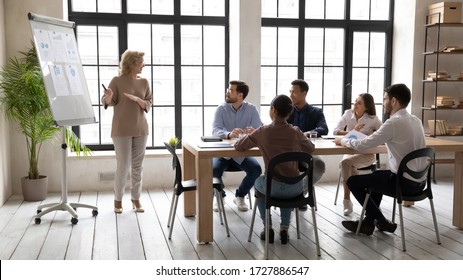 Smiling Mature 60 Year Old Businesswoman Flip Charts Presentation New Project In Boardroom At Company Meeting. Happy Coach Auditor Speaks With Diverse Colleagues About Business Using Board And Graphs.