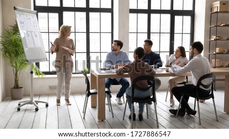 Smiling mature 60 year old businesswoman flip charts presentation new project in boardroom at company meeting. Happy coach auditor speaks with diverse colleagues about business using board and graphs.