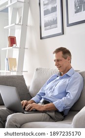 Smiling Mature 60s Older Senior Man Typing On Laptop Computer Tech Device Sitting On Sofa, Working Or E Learning Online, Using Banking Applications, Buying Insurance, Ordering Food On Website At Home.