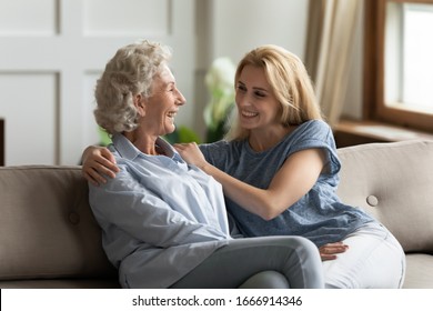 Smiling mature 70s mom and millennial girl child sit on couch in living room laugh talk on weekend together, happy senior mother and adult daughter rest on sofa at home, have fun chatting
