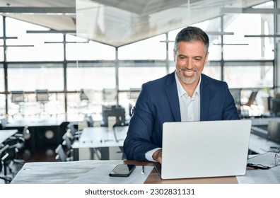 Smiling mature adult business man executive sitting at desk using laptop. Happy busy professional mid aged businessman ceo manager working on computer corporate technology in modern office. - Powered by Shutterstock