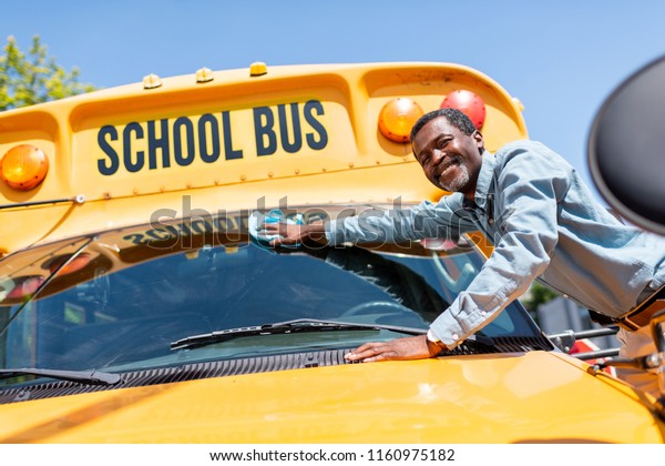 smiling mature african american\
bus driver wiping front window of school bus and looking at\
camera