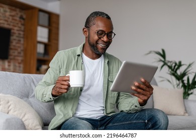 Smiling mature african american man in glasses drinking coffee from cup and watching video on tablet in living room interior. Chat in social networks remotely, new normal, app for working at home - Powered by Shutterstock