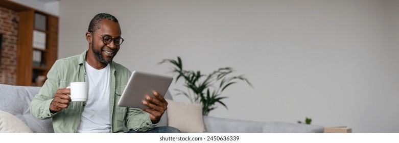 Smiling mature african american man in glasses drinking coffee from cup and watching video on tablet in living room interior. Chat in social networks remotely, app for working at home, copy space - Powered by Shutterstock