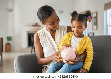 Smiling mature african american mother helping daughter sitting on lap putting money in piggy bank. Cute little black girl saving money by adding a coin in piggy bank with mother at home.  - Shutterstock ID 2126926490