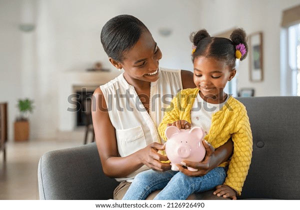 Smiling\
mature african american mother helping daughter sitting on lap\
putting money in piggy bank. Cute little black girl saving money by\
adding a coin in piggy bank with mother at home.\
