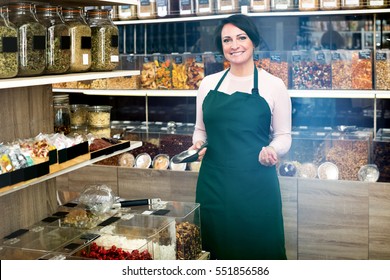 Smiling Mature Brunette Woman Wearing Apron And Selling Nuts And Dried Fruits In Organic Shop
