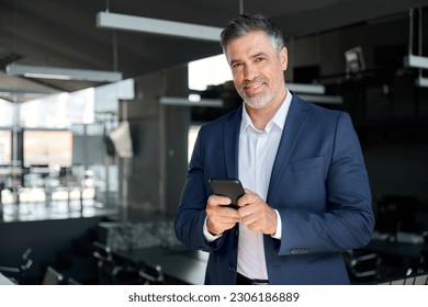 Smiling mature business man executive standing in office using cell phone. Happy mid aged businessman ceo professional manager holding smartphone satisfied with corporate mobile application solutions. - Powered by Shutterstock