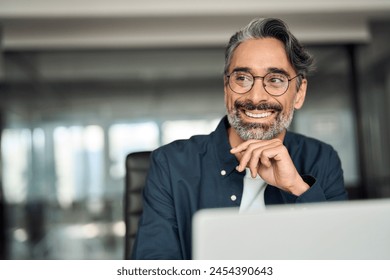 Smiling mature business man executive wearing shirt sitting at desk using laptop. Happy busy professional middle aged Indian businessman investor working on computer looking away in office. Copy space