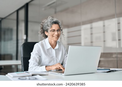 Smiling mature business woman executive working in office using laptop. Happy professional senior middle aged female worker wearing glasses and white shirt using computer sitting at work desk. - Powered by Shutterstock