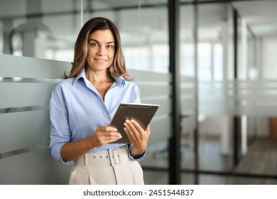 Smiling mature business woman executive, happy middle aged businesswoman entrepreneur, 40 years old company hr holding digital tablet looking at camera standing in office at work. Portrait.