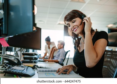 Smiling Mature Business Woman With Headset Working At Her Desk And Colleagues In Background.