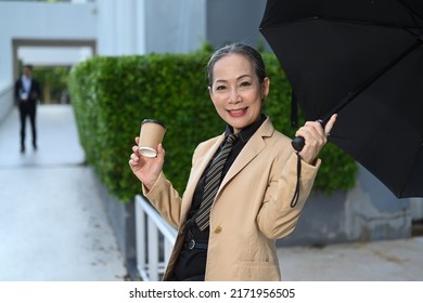 Smiling Mature Business Woman Holding Takeaway Coffee Cup And Sitting On Bench Under Umbrella At City Street