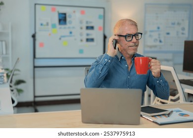 Smiling mature businessman drinking coffee and talking on mobile phone while working on laptop computer at office desk with whiteboard in background - Powered by Shutterstock