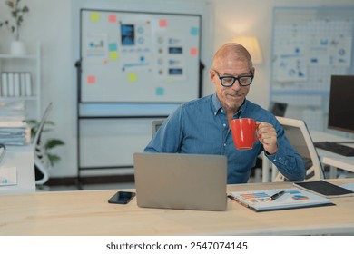 Smiling mature businessman drinking coffee while working on laptop computer at his desk in modern office, looking at screen and holding a red mug - Powered by Shutterstock