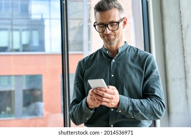 Smiling Mature Businessman Holding Smartphone Standing In Office. Middle Aged Manager Ceo Using Cell Phone Mobile Apps. Digital Technology Applications And Solutions For Business Corporate Development