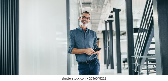 Lächelnder reifer Geschäftsmann, der ein Smartphone in einem Büro hält. Geschäftsmann, der die Kamera anschaut, während er allein auf einem modernen Arbeitsplatz steht. Erfahrener Geschäftsmann kommuniziert mit seinen Kunden.