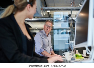 Smiling Mature Businessman Looking Through Glass Shield At Young Female Colleague In Office During Pandemic