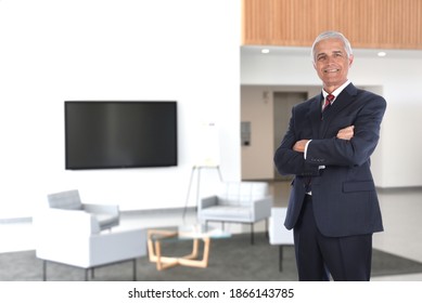 Smiling Mature Businessman Standing With Arms Folded In A Modern Office Building Lobby With Flat Screen TV.  Horizontal Format With A High Key Background.