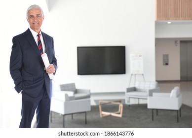 Smiling Mature Businessman Standing In A Modern Office Building Lobby With Flat Screen Monitor. He Is Holding A Rolled Up Document In His Hand. Horizontal Format With A High Key Background.