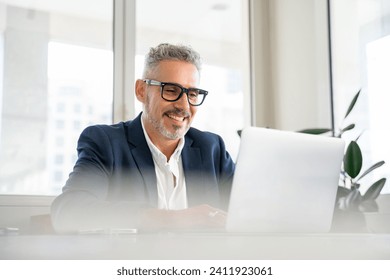 Smiling mature businessman wearing formal suit and glasses using laptop for office work, male employee looking at the screen, reading news, checking mail - Powered by Shutterstock