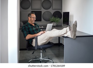 Smiling mature businessman working online with a laptop while sitting with his feet up on his desk in an office cubicle