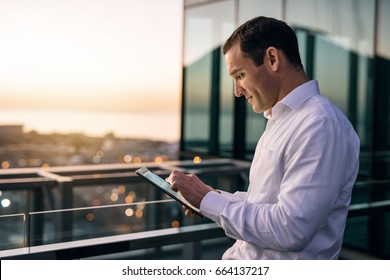 Smiling mature businessman working online with a digital tablet while standing outside on an office building balcony overlooking the city at dusk