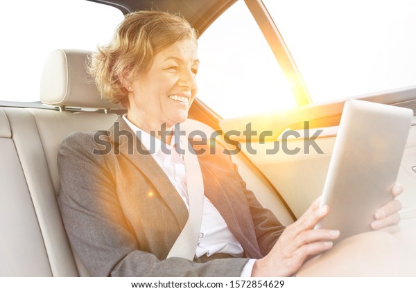 Smiling Mature businesswoman\
using digital tablet while sitting inside the car with yellow lens\
flare