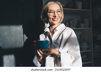 Smiling mature caucasian businesswoman with cup of tea or coffee looking away at dark home art studio full of ceramic products. Small business and entrepreneurship. Modern successful woman