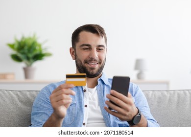 Smiling mature caucasian man shopaholic pays for goods with credit card and smartphone in living room interior. Black friday, online shopping, sale due covid-19 lockdown self-isolation, empty space
