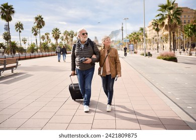 Smiling mature Caucasian tourist couple walking embraced carefree with their luggage looking to the side. Happy husband and wife enjoying their outdoor pensioner vacation in a winter sunny day - Powered by Shutterstock