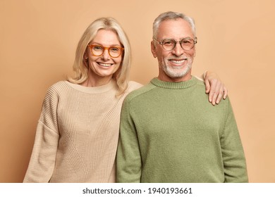 Smiling Mature Couple Embrace Look Gladfully At Camera Pose For Family Portrait Happy Children Came To Visit Them Wear Transparent Glasses Casual Jumpers Isolated Over Brown Background. Age Concept