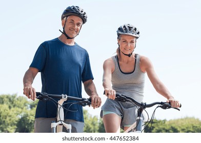 Smiling Mature Couple On A Mountain Bike Wearing Sports Clothes And Looking At Camera. Senior Couple Riding A Bike Together. Athletic Couple Enjoying Cycle Ride And Looking At Camera.
