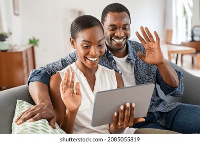 Smiling mature couple waving hands during video call using digital tablet. Middle aged black man and beautiful woman sitting on couch together doing video conference with family or friends with laptop