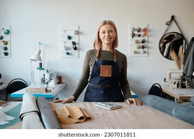 Smiling mature craftswoman in apron looking at camera in workshop or studio while standing by table with various leather and suede textile - Powered by Shutterstock