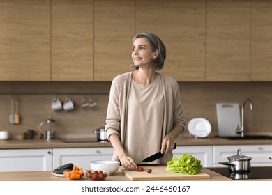 Smiling mature dreamy woman stand at table, cutting fresh vegetables for salad, looking away, smile, planning dinner menu, use healthy ingredients, enjoy cooking process in modern kitchen. Culinary