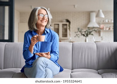 Smiling mature elder 60s woman sitting relaxing with cup of tea, coffee. Senior mid age stylish look woman with eyeglasses portrait with cup looking away at modern home. - Powered by Shutterstock