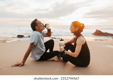 Smiling mature european couple in sportswear, rest from workout, drink bottle of water together in morning, sit on sea beach, outdoor. Relationship, sports, fitness and body care - Powered by Shutterstock