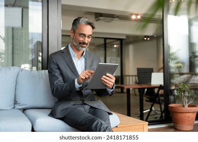 Smiling mature European Latin businessman holding tablet gadget sitting in office with copy space. Middle aged man manager using digital device app. Technology application and solutions for business. - Powered by Shutterstock