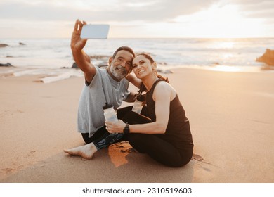Smiling mature european woman and man in sportswear rest from training together, drink bottle of water, take selfie on phone in morning on sea beach, outdoor. Sports blog, fitness and body care - Shutterstock ID 2310519603